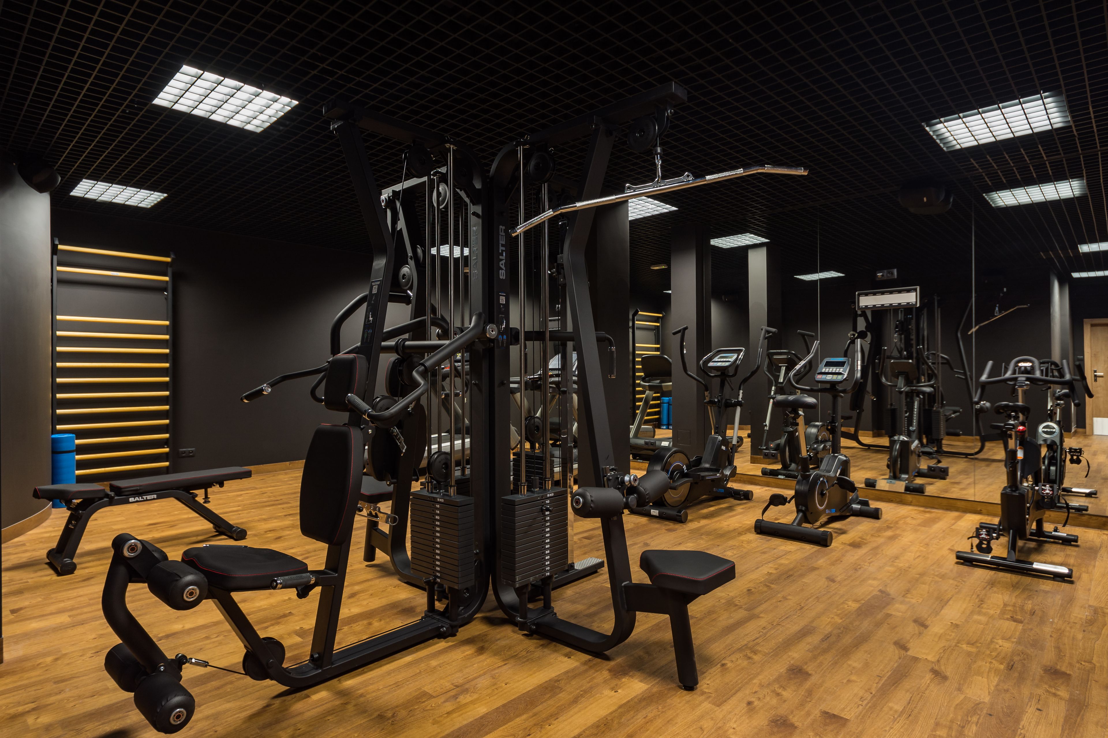 A gym with various exercise machines, wooden flooring, and a high ceiling with grid lighting.