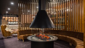 A modern lounge area with a central fire feature, cushioned seating, and a bar in the background.