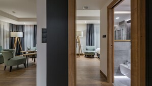 A modern hotel room with a wooden floor, a dining area with a table and chairs, a sofa, and a view of the bathroom with a bathtub and sink.