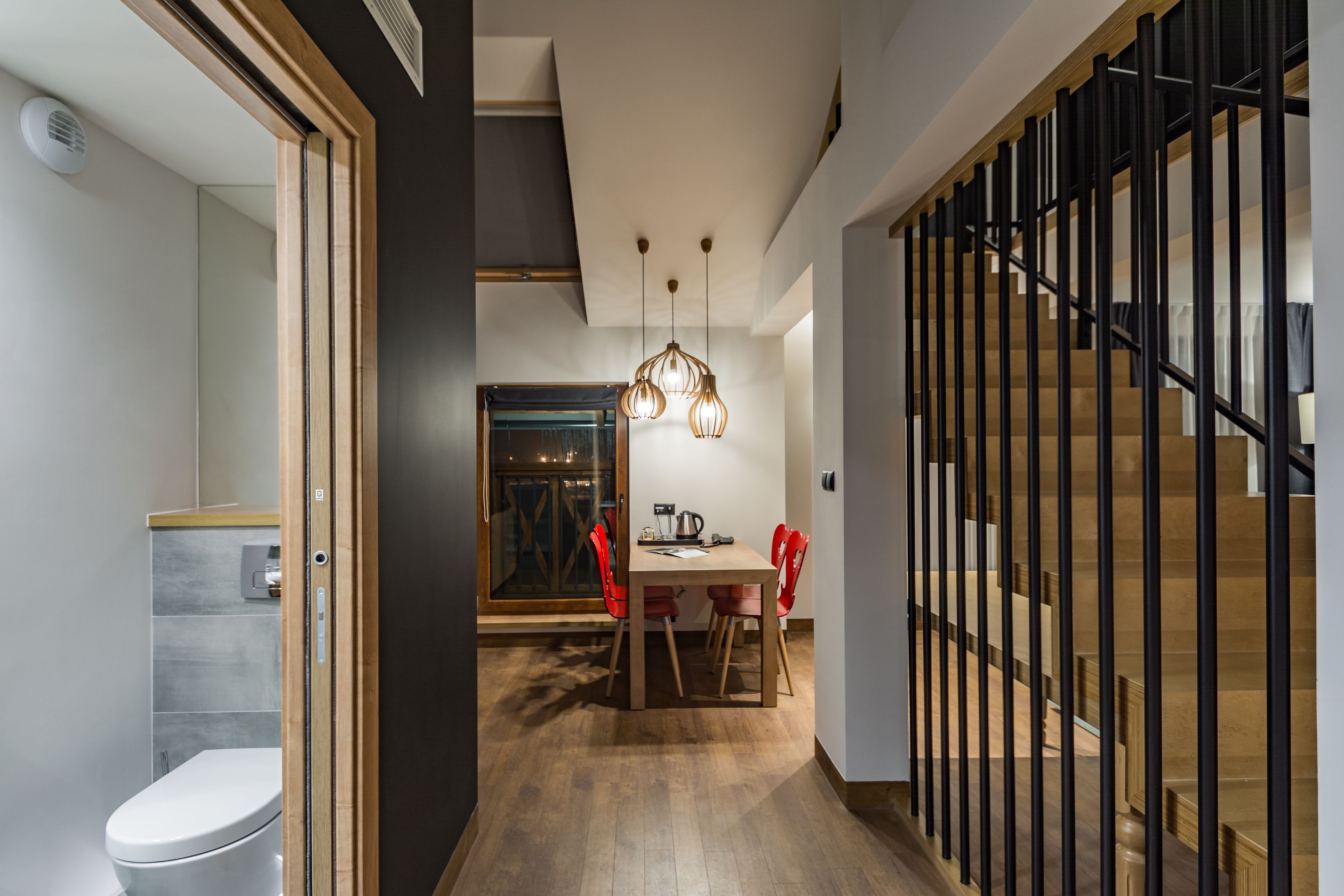 Un intérieur moderne avec un escalier en bois, un coin repas avec des chaises rouges et une cuisine visible à travers une porte ouverte.