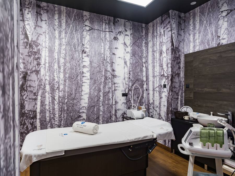 A room with a birch forest wallpaper, a massage table, a sink, and a treatment cart.