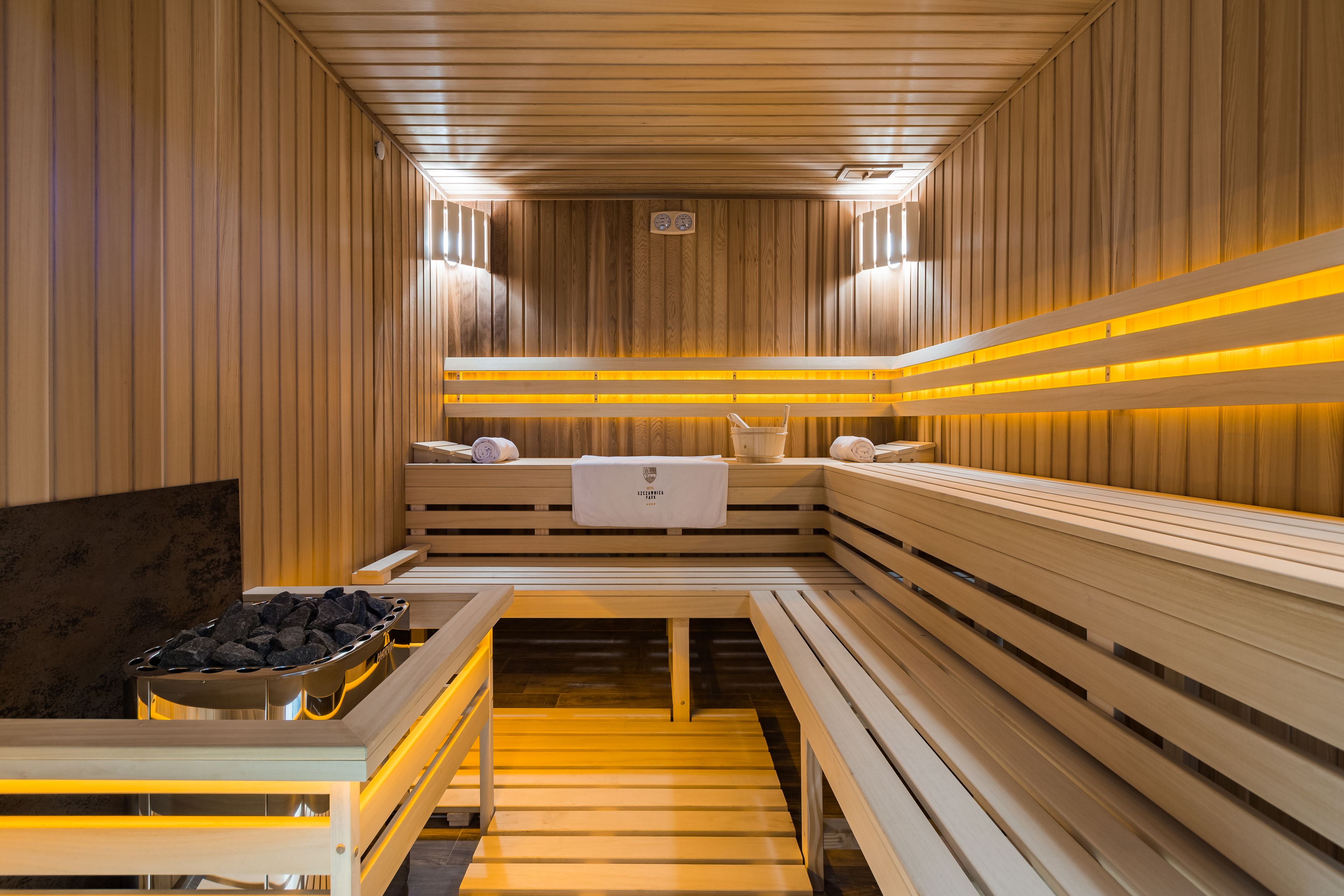 Uma sauna com bancos de madeira e uma mesa central, equipada com toalhas e um cesto de pedras.