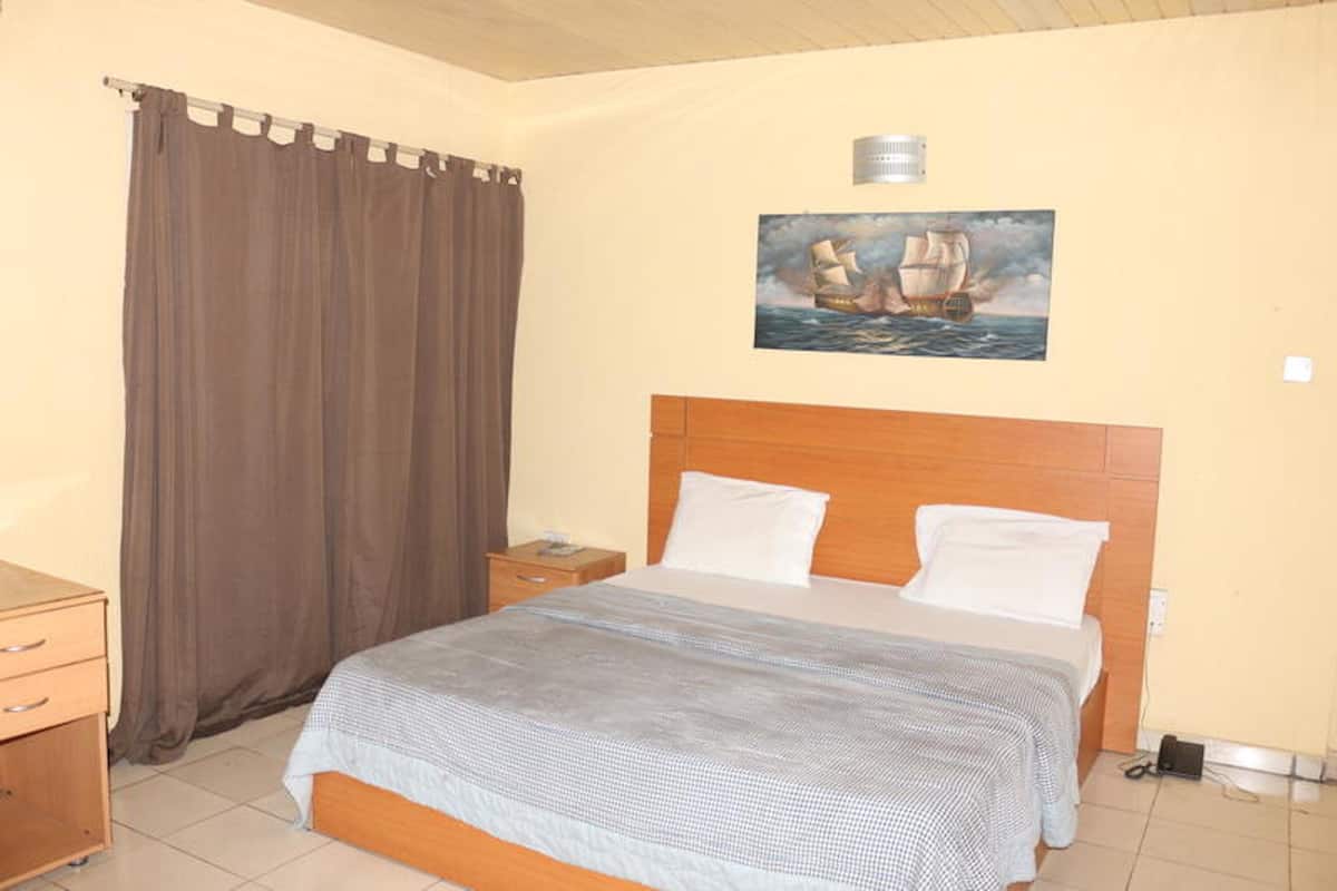 A bedroom with a bed, a nightstand, a wooden headboard, a painting of a ship on the wall, and a curtain.