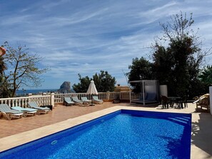 A swimming pool with lounge chairs and umbrellas, surrounded by a fence and trees, with a view of a mountain and cityscape.