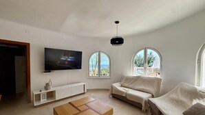 A modern living room with a flat-screen TV, a wooden coffee table, a beige sofa, and two large windows offering a view of the outdoors. - Bellavista - Villa with panoramic sea views and large terraces (Calpe / Calp)