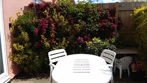 A patio with a round table and four chairs, surrounded by lush greenery and flowering plants. - Number Nine New Esplanade is a quiet coastal snug with a flat walk to the shore (Paignton)