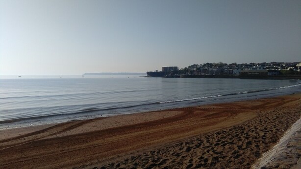 A beach with a clear view of the sea and a distant shoreline with buildings. - Number Nine New Esplanade is a quiet coastal snug with a flat walk to the shore (Paignton)