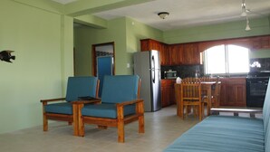 A living room with two blue chairs, a wooden dining table, and a kitchen area with wooden cabinets and a refrigerator.