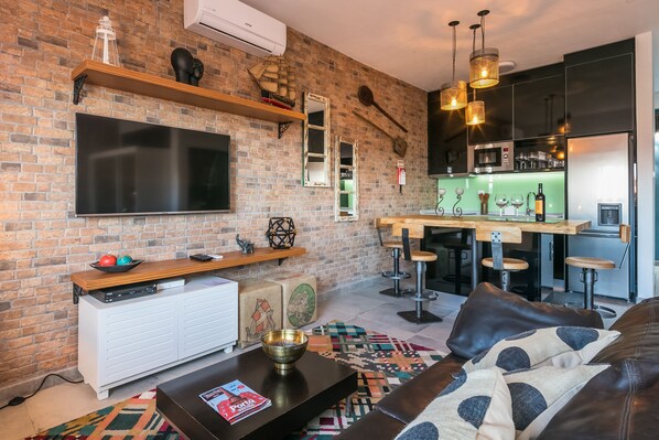 A modern living room with a brick wall, a flat-screen TV, a wooden shelf, a kitchen area with bar stools, and a leather sofa.