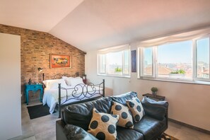 A bedroom with a brick wall, a bed, a black leather sofa, and a large window with a city view.