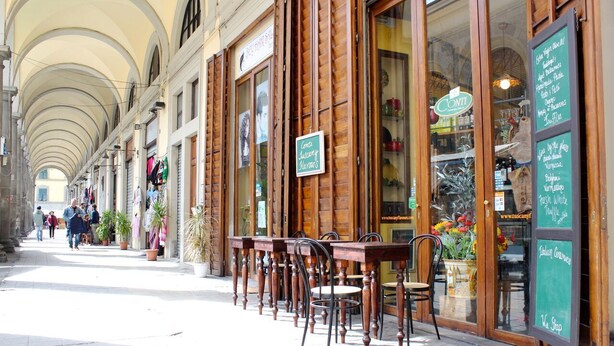 A row of wooden tables and chairs outside a building with large windows and a signboard. - APARTMENT CLOSE TO THE CATHEDRAL & RAILWAY STATION,WI-FI (Firenze)