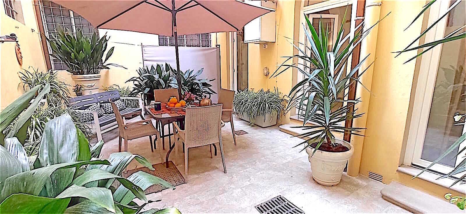 A patio area with a dining table, chairs, and a large umbrella. There are potted plants and a window with a view.