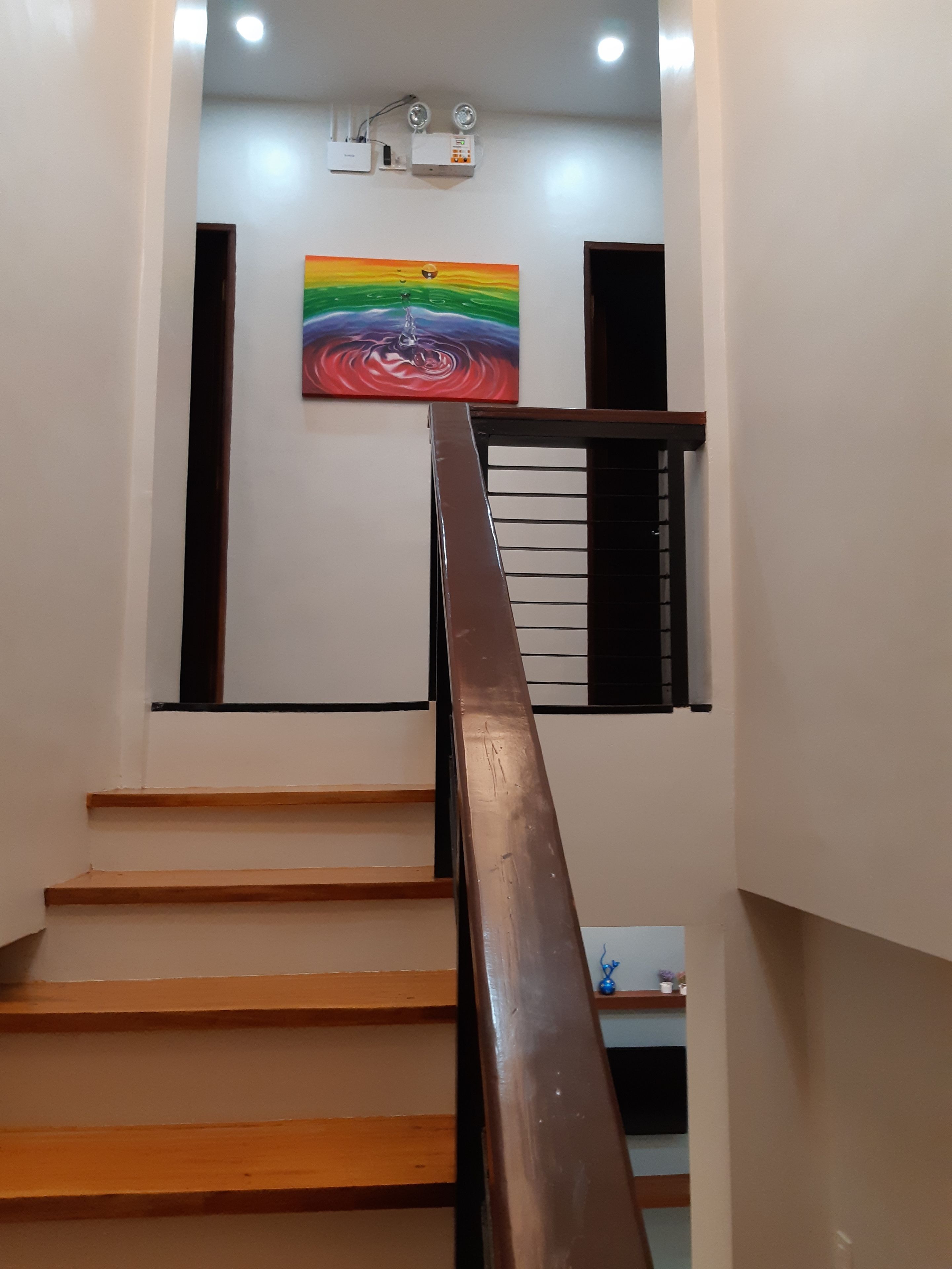 Un escalier avec des marches en bois et une rampe métallique, menant à un palier orné d’un tableau coloré et où sont fixés deux appareils de climatisation au mur.
