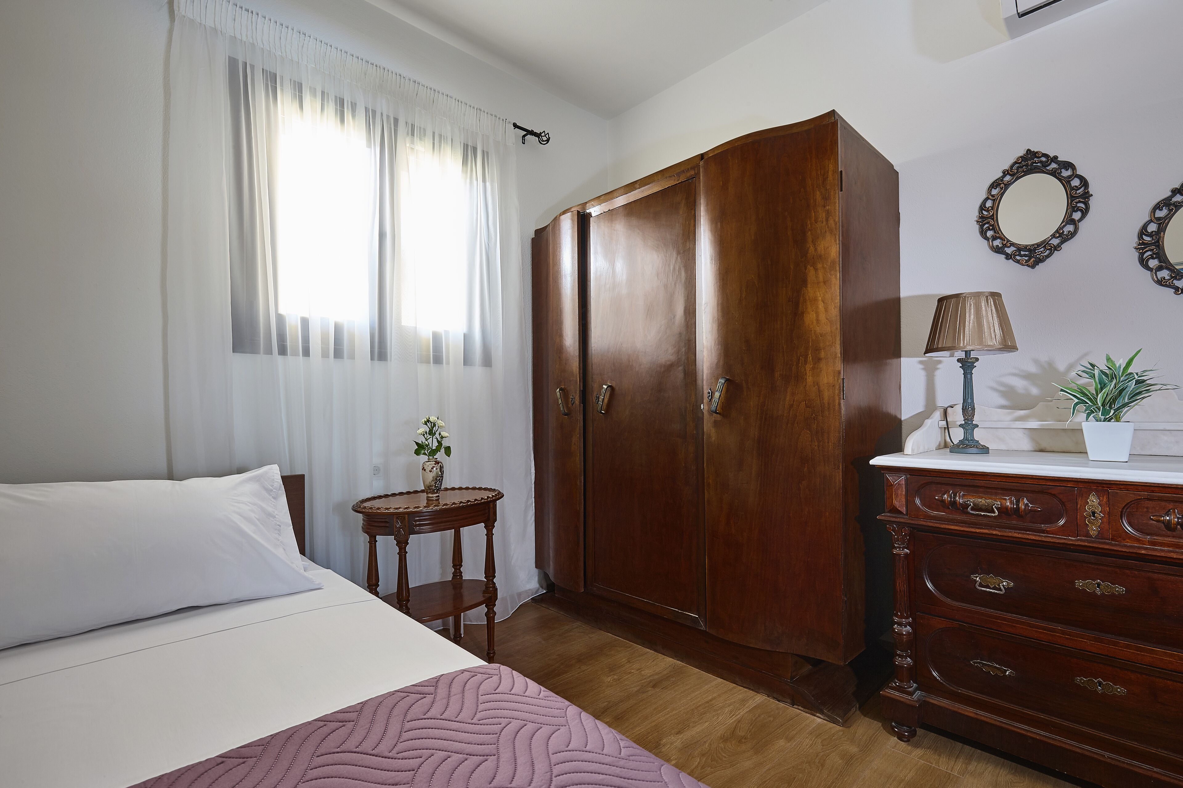 A bedroom with a bed, a wooden wardrobe, a small table with a vase, and a dresser with drawers.