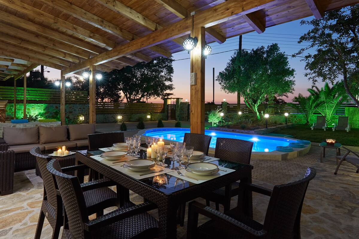 An outdoor dining area with a pool, wooden pergola, and stone flooring.