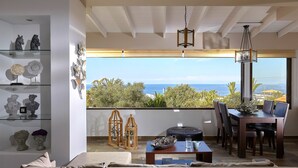 A modern living room with a large window overlooking a scenic ocean view, a dining area with a table and chairs, and a bookshelf with decorative items. - Feronia Villas (Hersonissos)