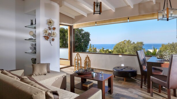 A modern living room with a sofa, coffee table, and chairs, a balcony with a view of the sea, and decorative items on shelves. - Feronia Villas (Hersonissos)