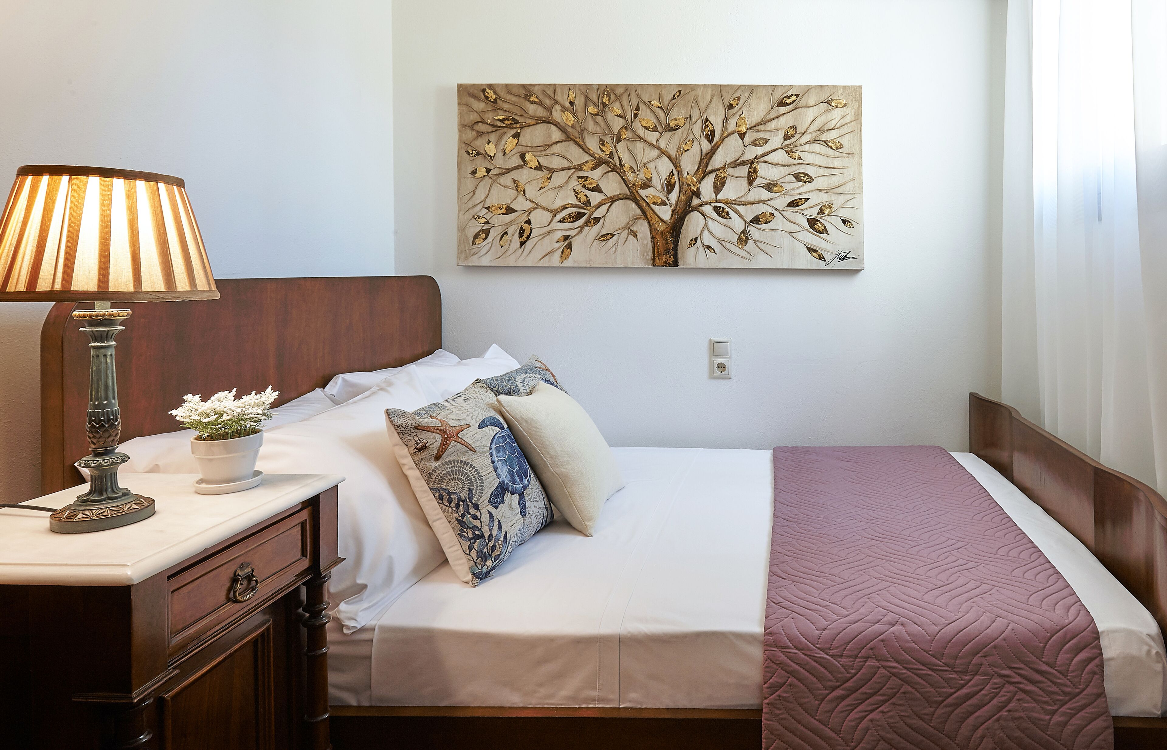 A neatly made bed with a pink quilt, a wall-mounted painting of a tree, a bedside lamp, and a nightstand with a lamp and a small potted plant.