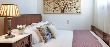 A neatly made bed with a pink quilt, a wall-mounted painting of a tree, a bedside lamp, and a nightstand with a lamp and a small potted plant.