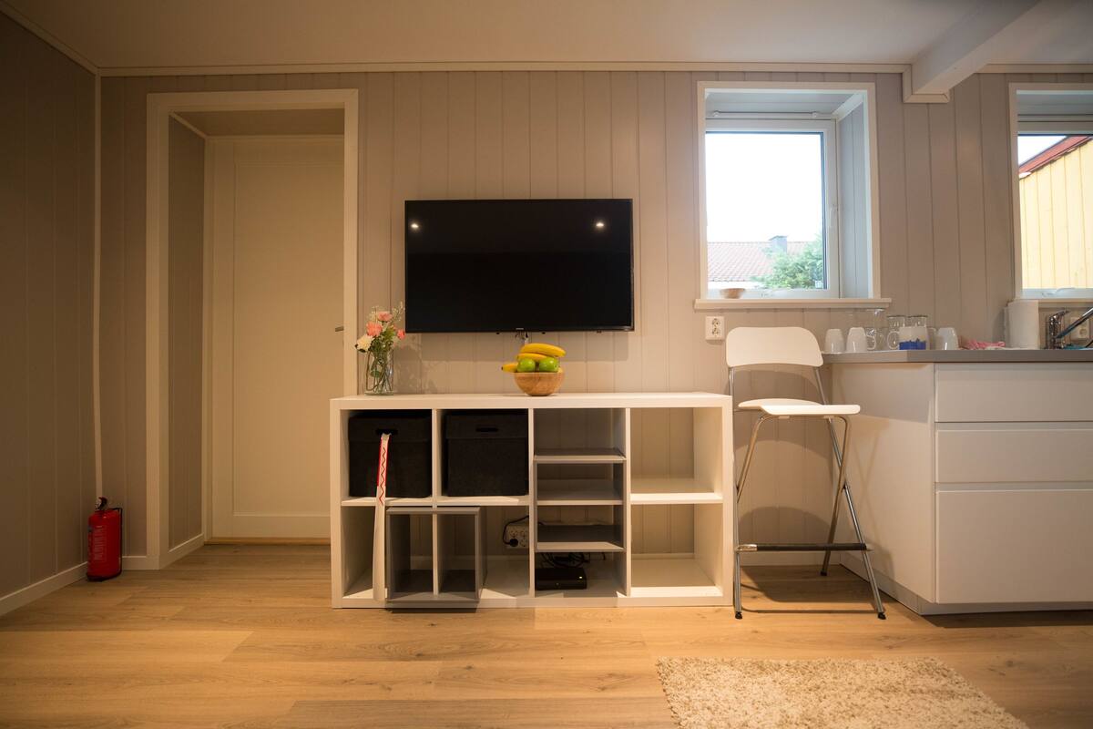 Un salon moderne avec un téléviseur à écran plat fixé au mur, un meuble de rangement blanc, une petite table blanche et une fenêtre donnant sur l’extérieur.