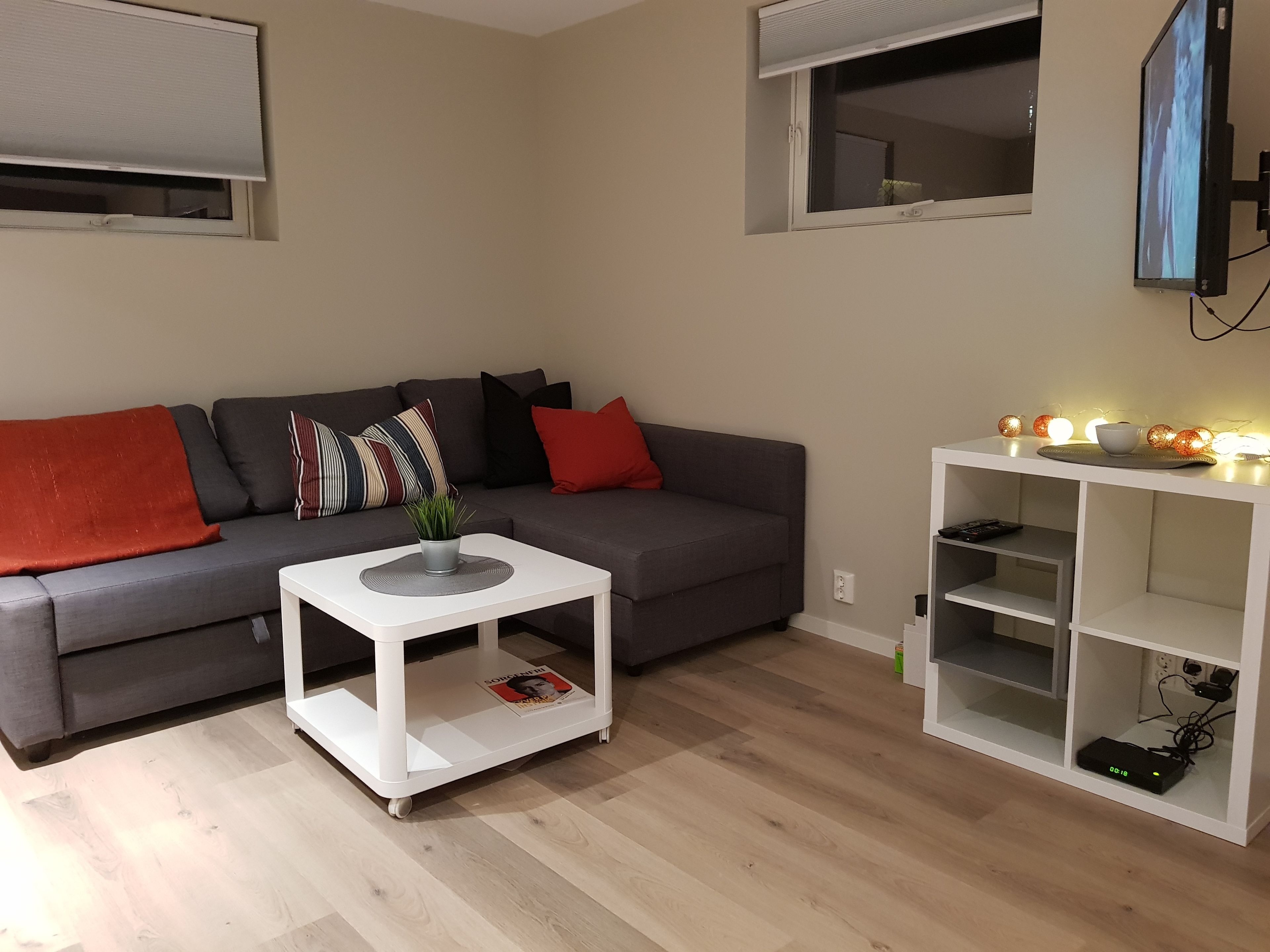 Una sala de estar moderna con un sofá gris, almohadas rojas y a rayas, una mesa de centro blanca con una revista, y un estante blanco con luces decorativas.