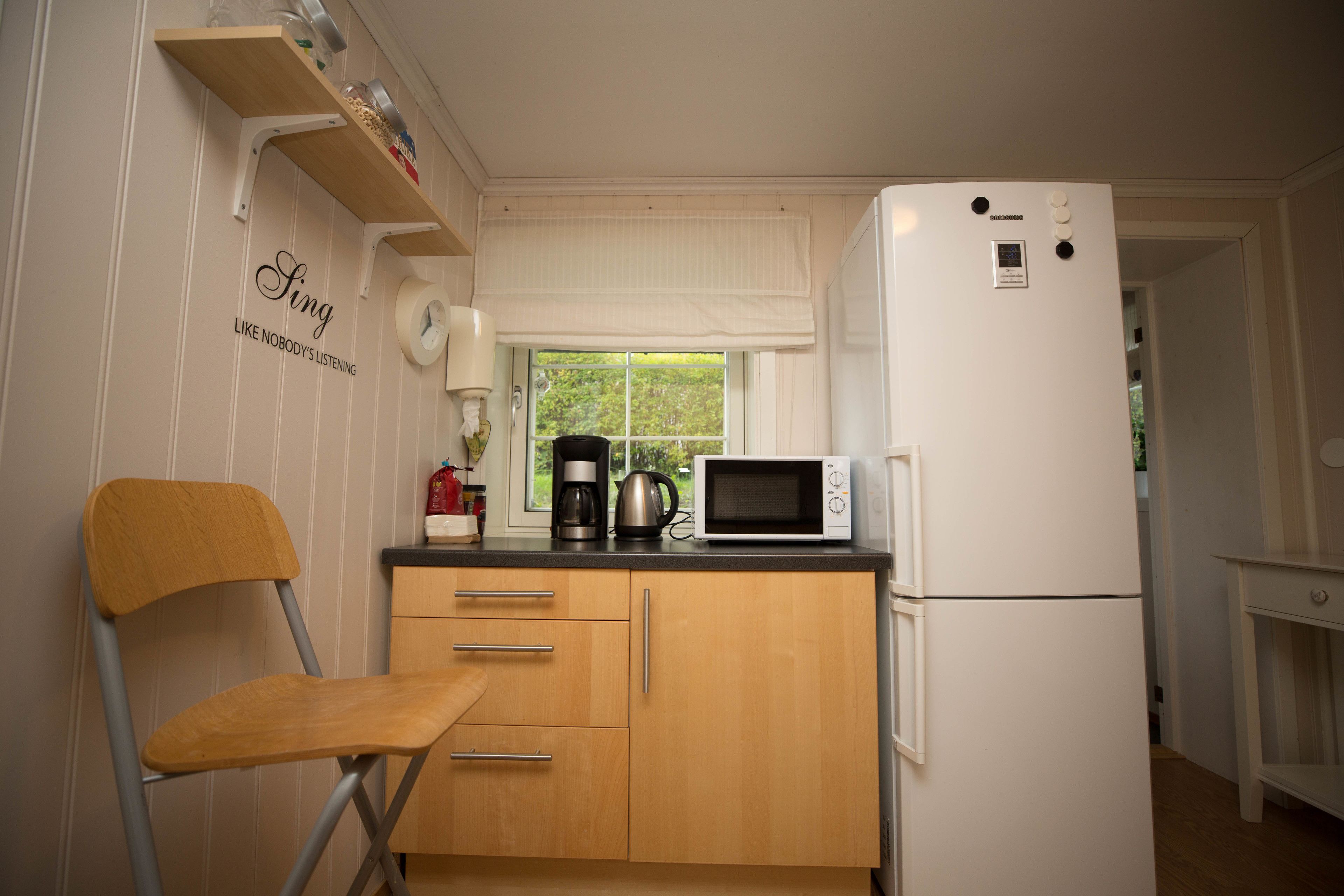 Una cocina compacta con microondas, cafetera y refrigerador. Hay una silla de madera y una ventana con cortinas.