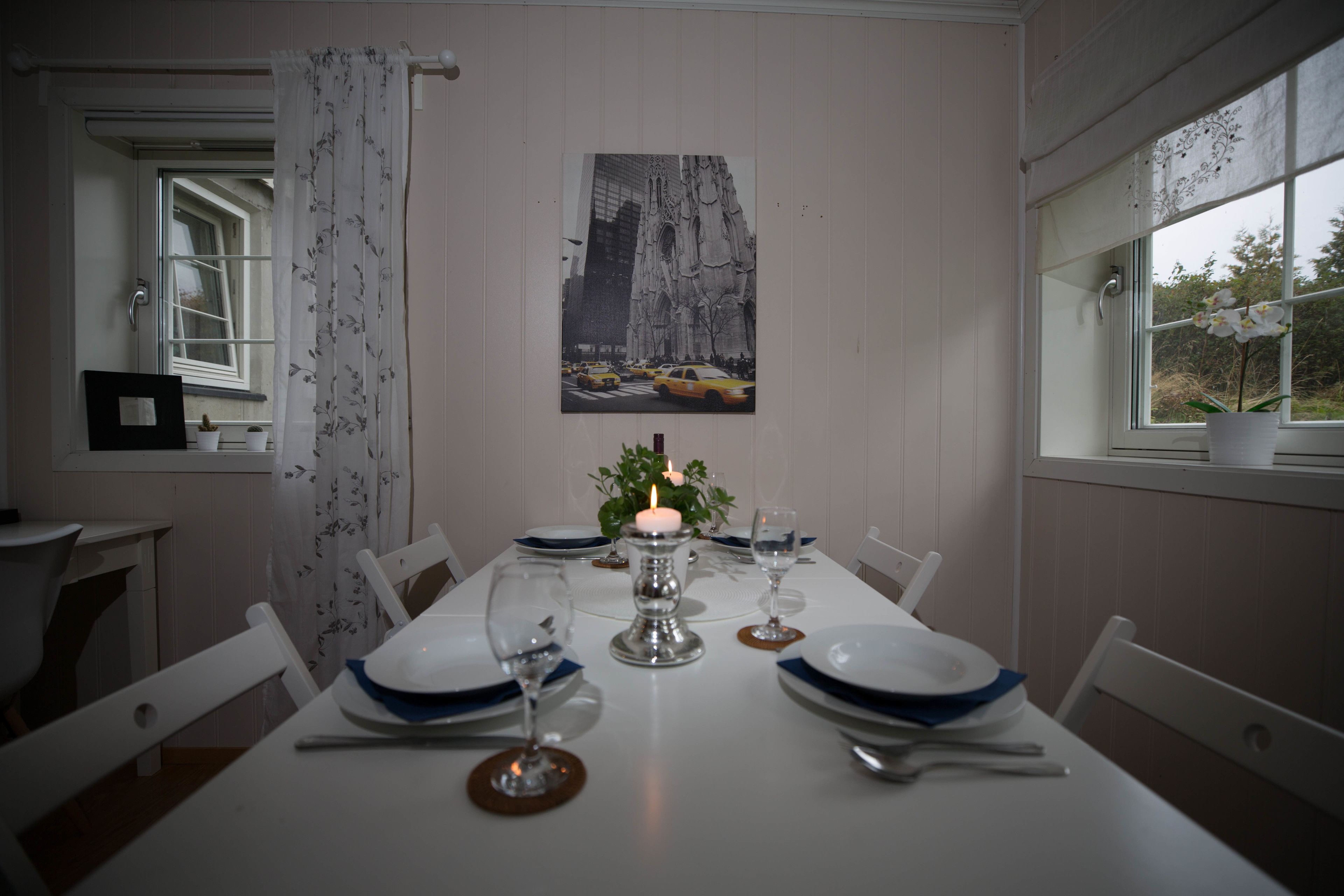 A dining table set with plates, cutlery, and a candle, with a cityscape photo on the wall and a window with a view of trees.