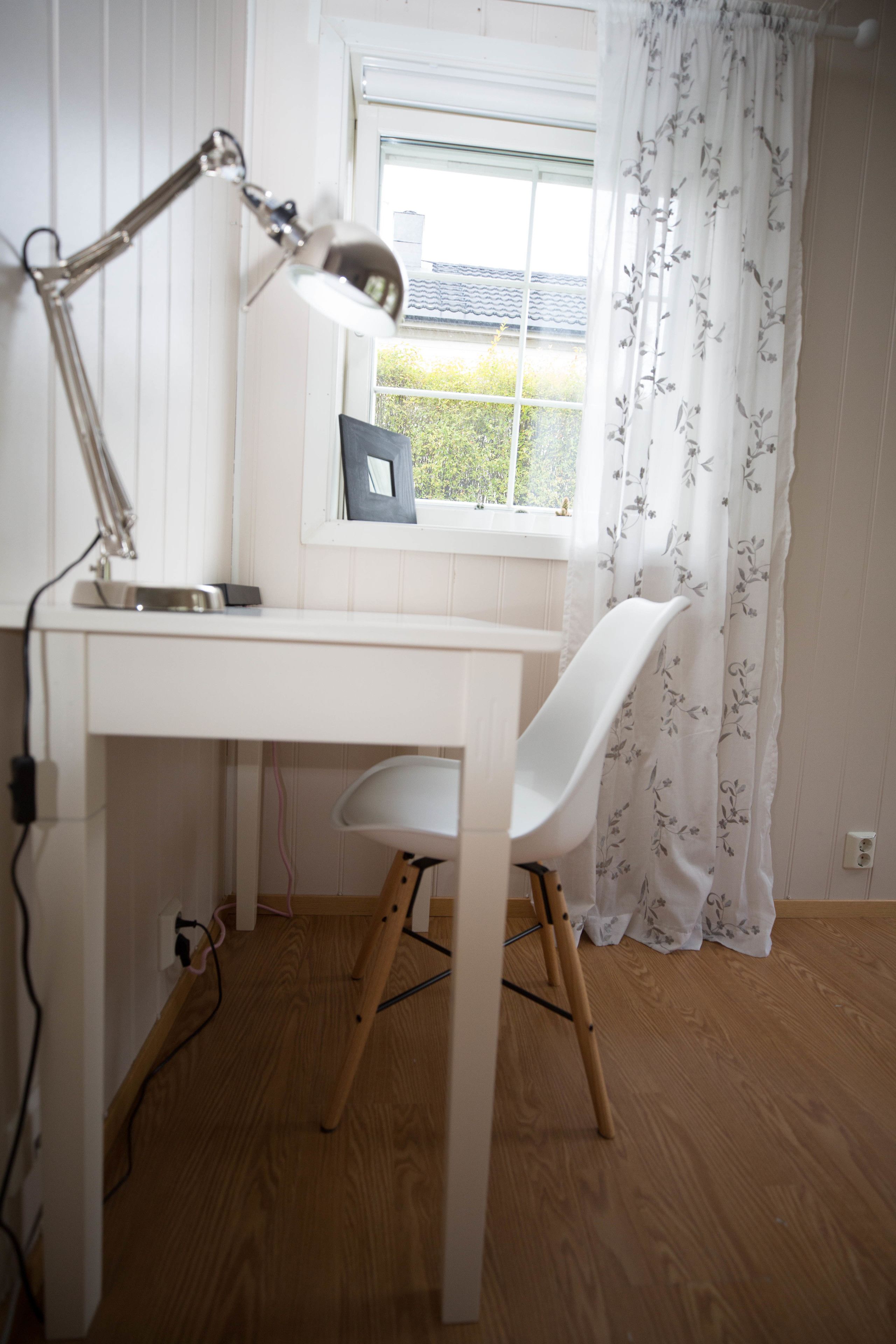 A small, white desk with a lamp, a chair, and a window with a view of greenery.