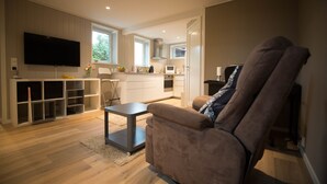 A modern living room with a flat-screen TV, a comfortable sofa, a small coffee table, and a kitchen area with a microwave and oven.