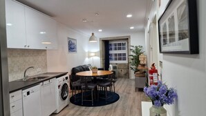 A compact kitchen with a sink, dishwasher, and washing machine. There is a dining area with a round table and chairs, and a small side table with a vase of flowers. - Merc Porto Ribeira View (Porto)