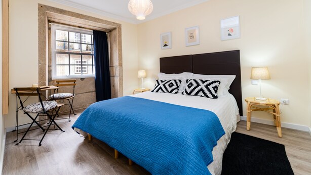 A bedroom with a bed, two bedside tables with lamps, a window with curtains, a small table with chairs, and artwork on the wall. - Merc Porto Ribeira View (Porto)