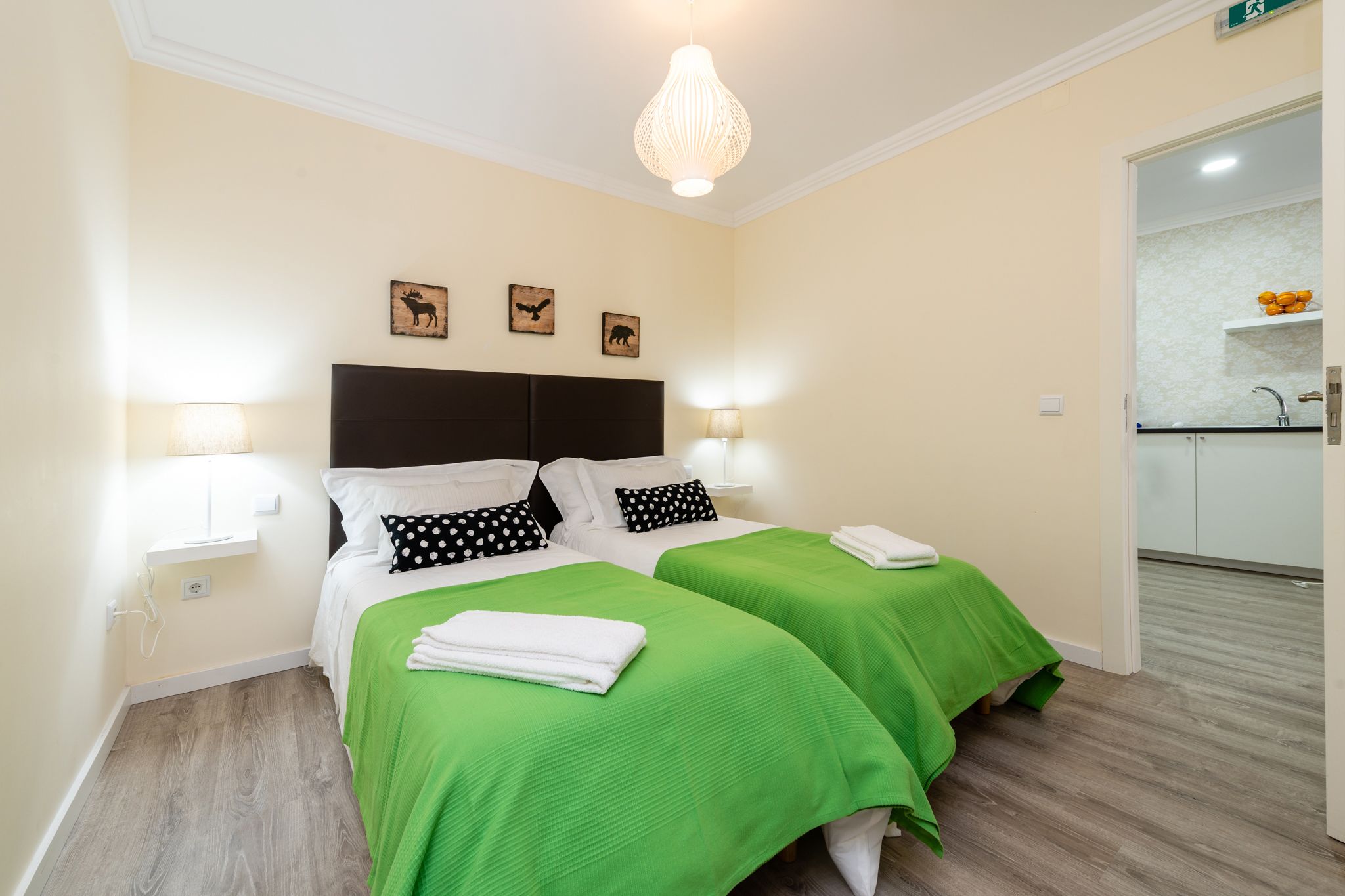 A double room with two beds, each with green bedding and white pillows, a dark headboard, bedside lamps, and wall art.