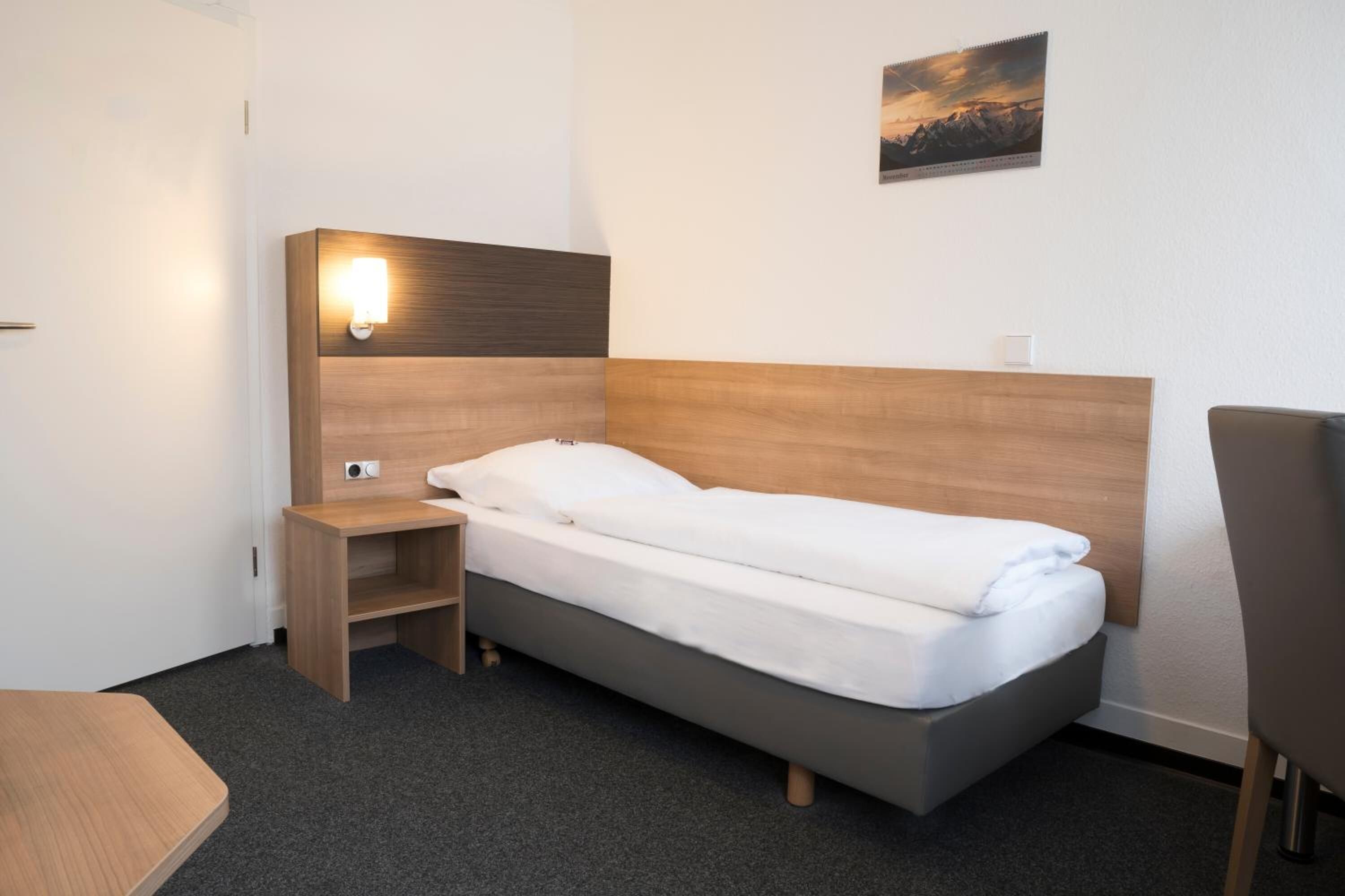 A single bed with a wooden headboard and a nightstand, a small table, and a chair in a hotel room.