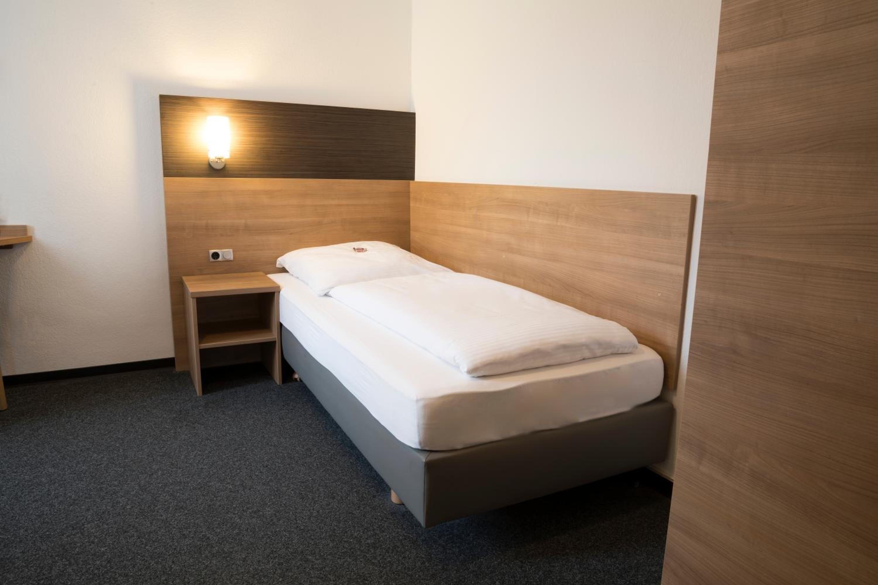 A small, neatly arranged hotel room with a single bed, a nightstand, and a wall-mounted light.