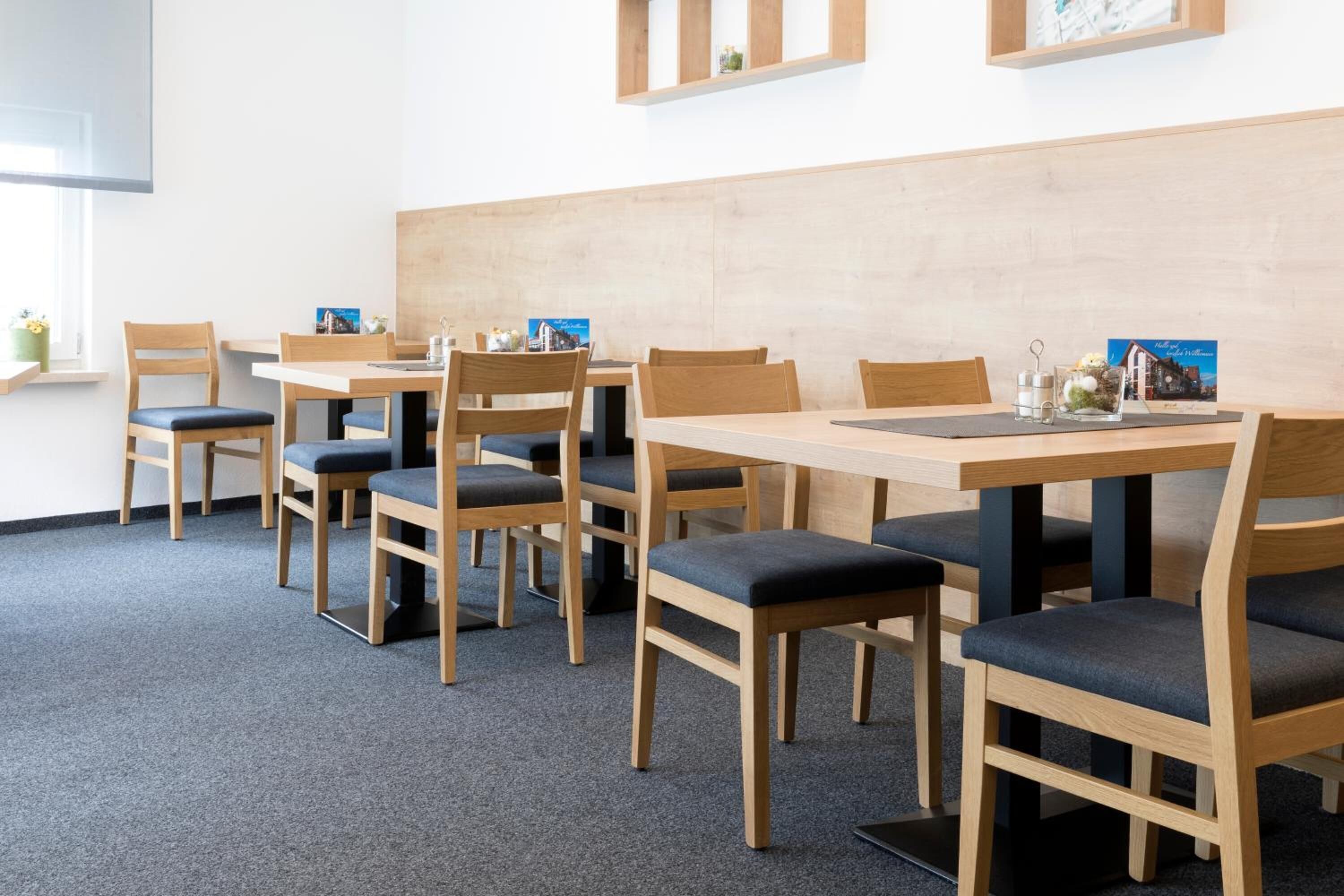 Un espace repas avec des tables et des chaises en bois, un sol recouvert d’une moquette et une étagère fixée au mur, ornée de petits objets décoratifs.