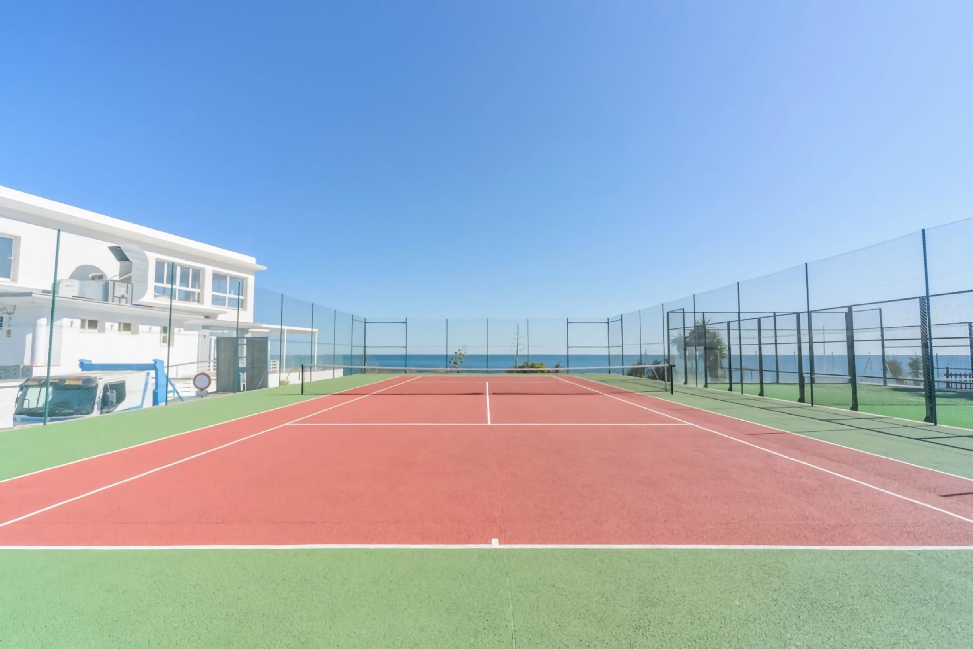 A tennis court with a net, surrounded by a fence, and a building with a blue door and windows in the background.
