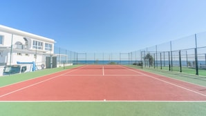 Una cancha de tenis con una red, rodeada por una valla, y un edificio con una puerta y ventanas azules al fondo.