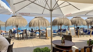 Un café frente a la playa con mesas al aire libre, sombrillas y vistas al mar.