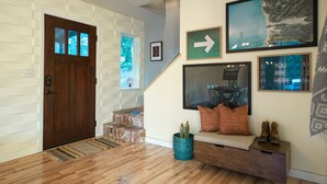 Un interior moderno con una puerta de madera, una escalera, una zona de estar con cojines, un televisor montado en la pared y una obra de arte enmarcada.