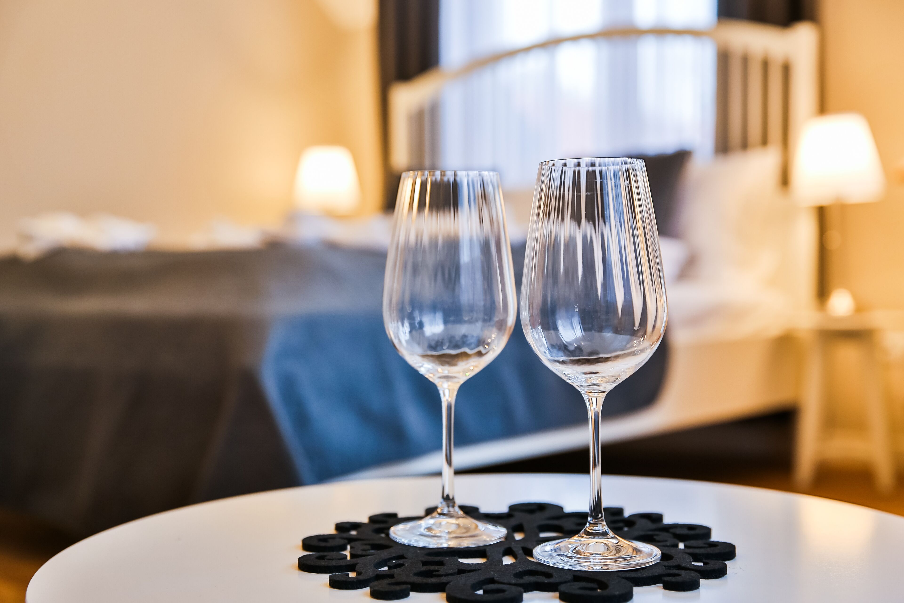 Deux verres à vin vides sont posés sur une table blanche, accompagnés d’un sous-verre noir en forme de puzzle, dans une chambre d’hôtel équipée d’un lit et d’une table de chevet avec une lampe.