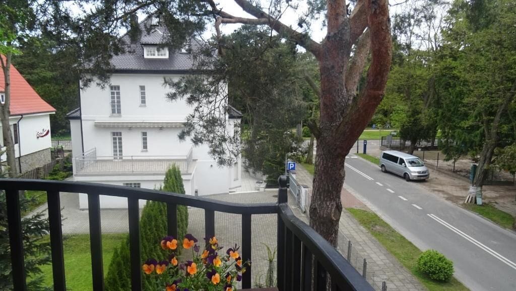 Een wit, twee verdiepingen hoog huis met een zichtbaar parkeersbord, een boom op de voorgrond en een weg met een geparkeerde auto.
