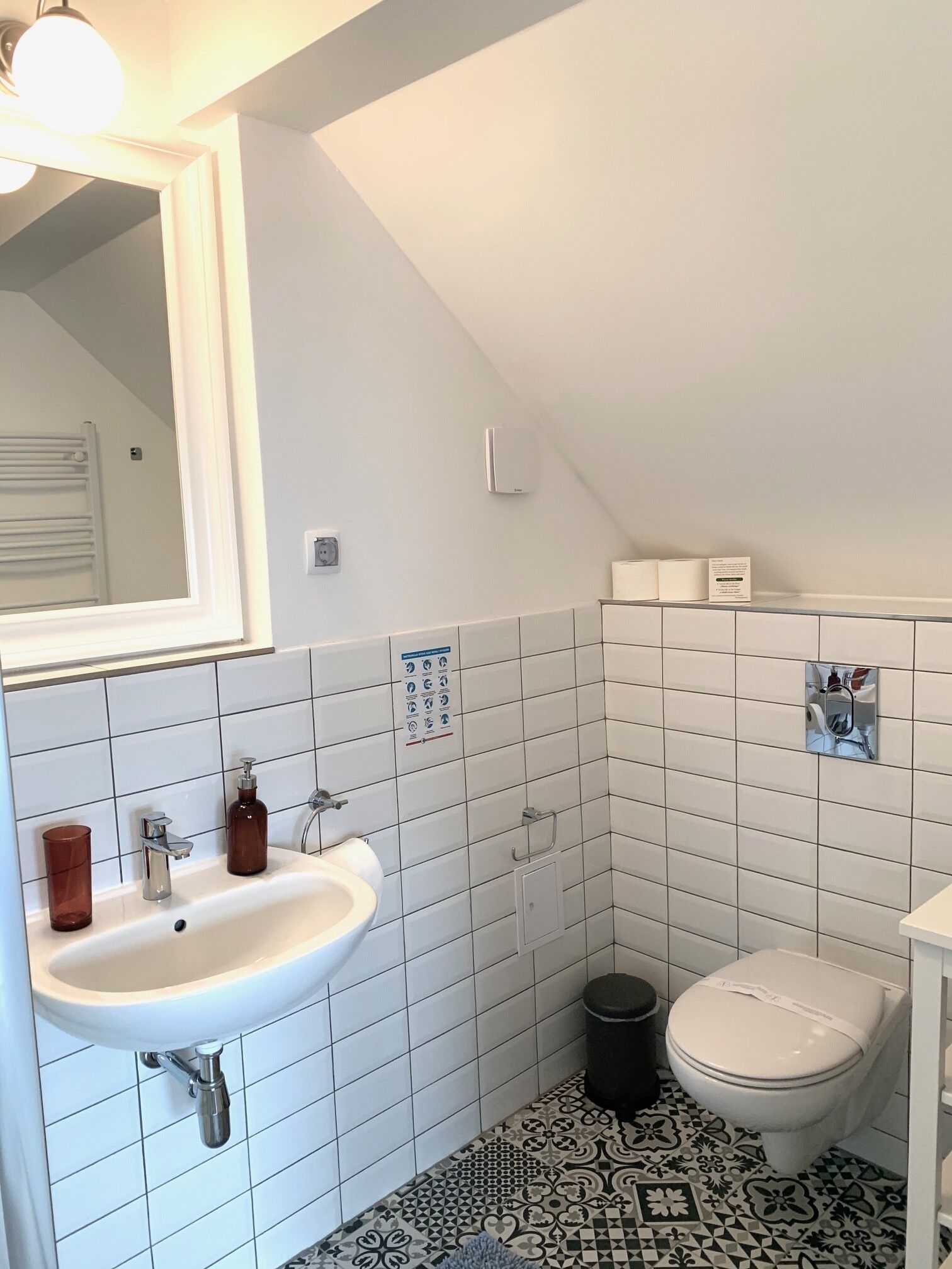 A bathroom with a white sink, a toilet, and a mirror. There are white tiles on the walls and floor, and a patterned tile design near the toilet.