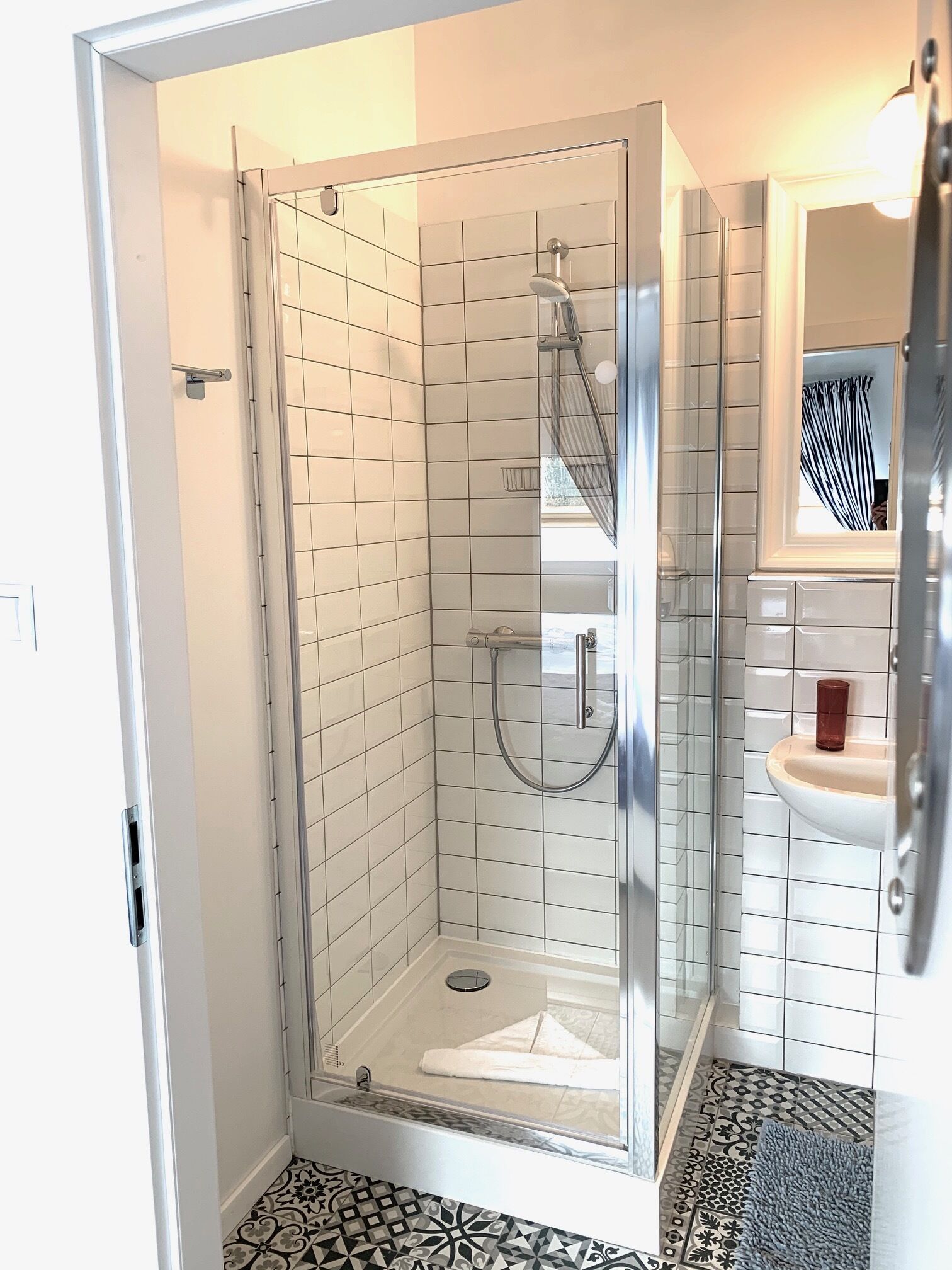 Un baño moderno con ducha acristalada, paredes cubiertas de azulejos blancos, un lavabo con espejo y baldosas de piso estampadas.
