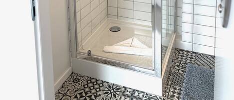 Un baño moderno con ducha acristalada, paredes cubiertas de azulejos blancos, un lavabo con espejo y baldosas de piso estampadas.