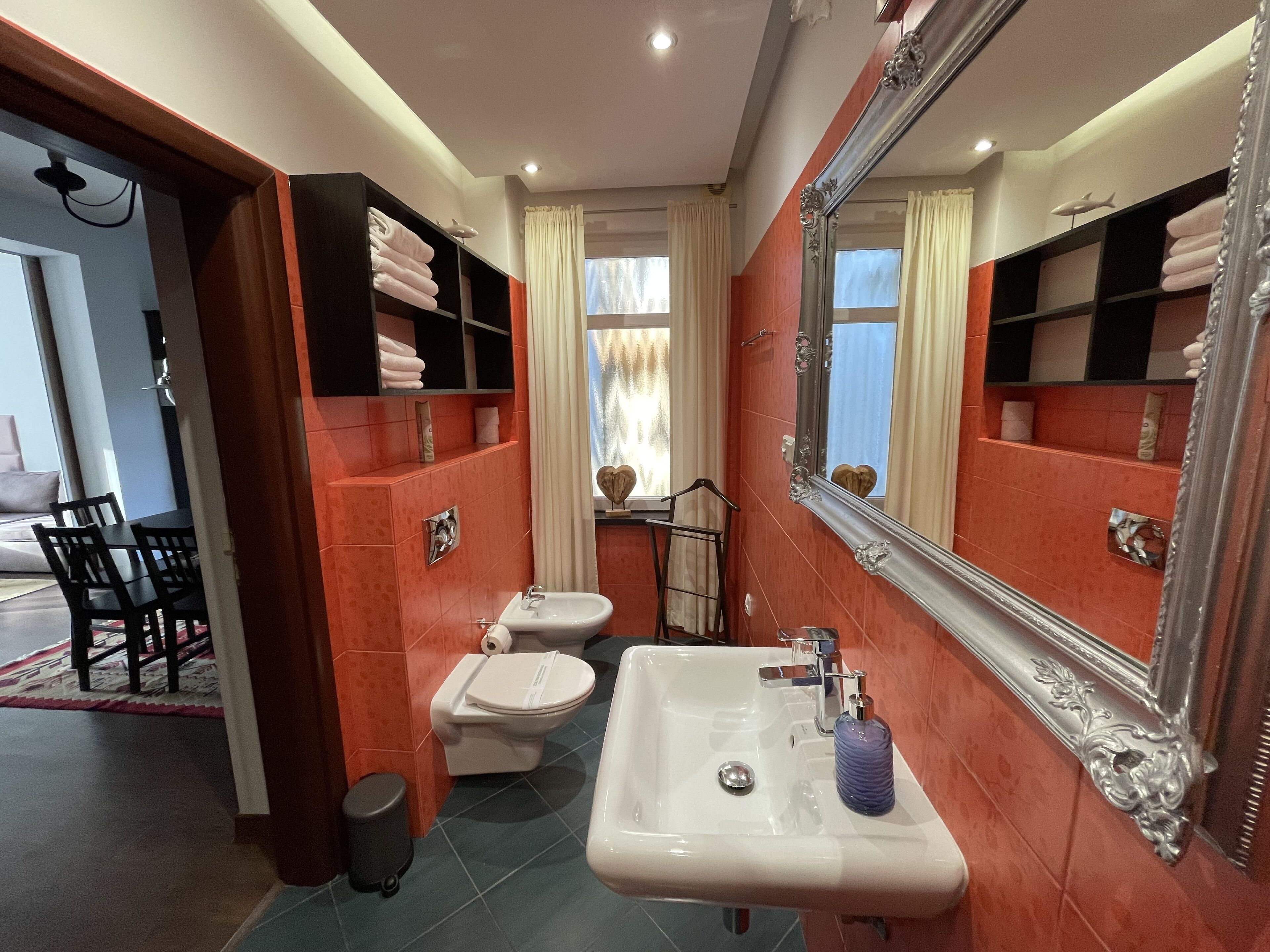 Ein Badezimmer mit Toilette, Waschbecken und einem großen Spiegel. Handtücher liegen auf Regalen und ein Fenster ist mit Vorhängen versehen.