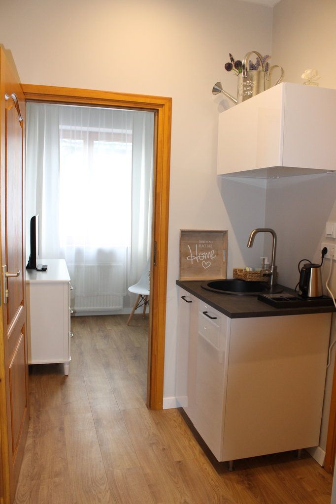 A compact kitchen with a white countertop, a sink, and a small cabinet. There is a framed sign on the wall and a window with sheer curtains.