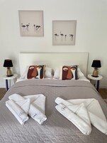 Una cama bien tendida con toallas, dos mesitas de noche con lámparas y dos obras de arte con flamencos.