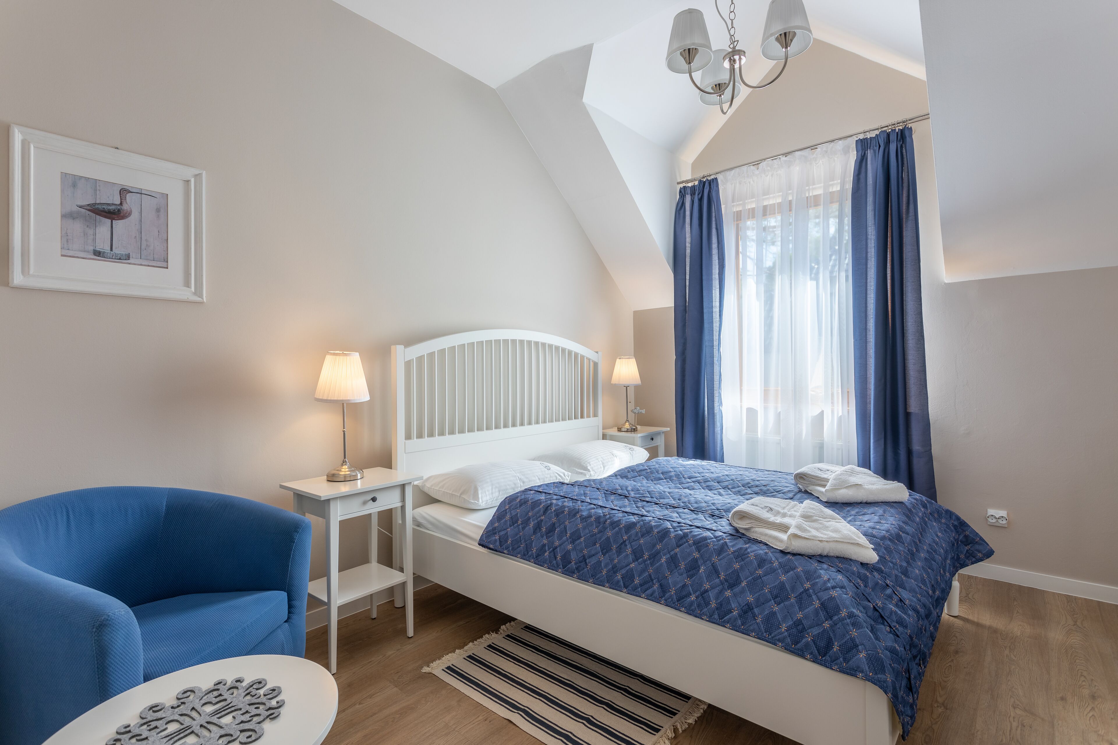 Un dormitorio con cama blanca, cortinas azules, mesita de noche blanca, una silla azul y una pequeña mesa con adornos.