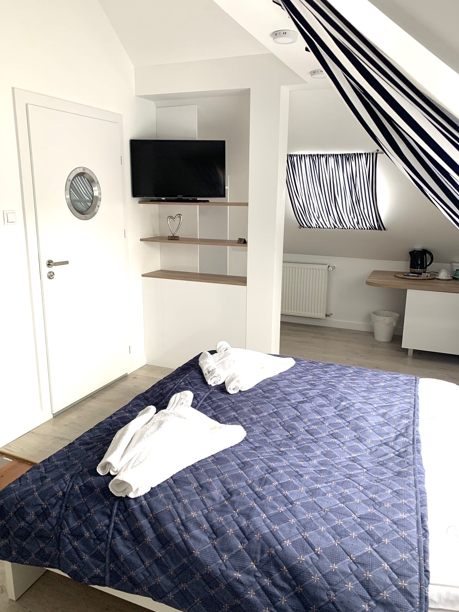 Una habitación de hotel con una cama, un televisor montado en la pared, un pequeño estante con un objeto con forma de corazón y una cortina a rayas.