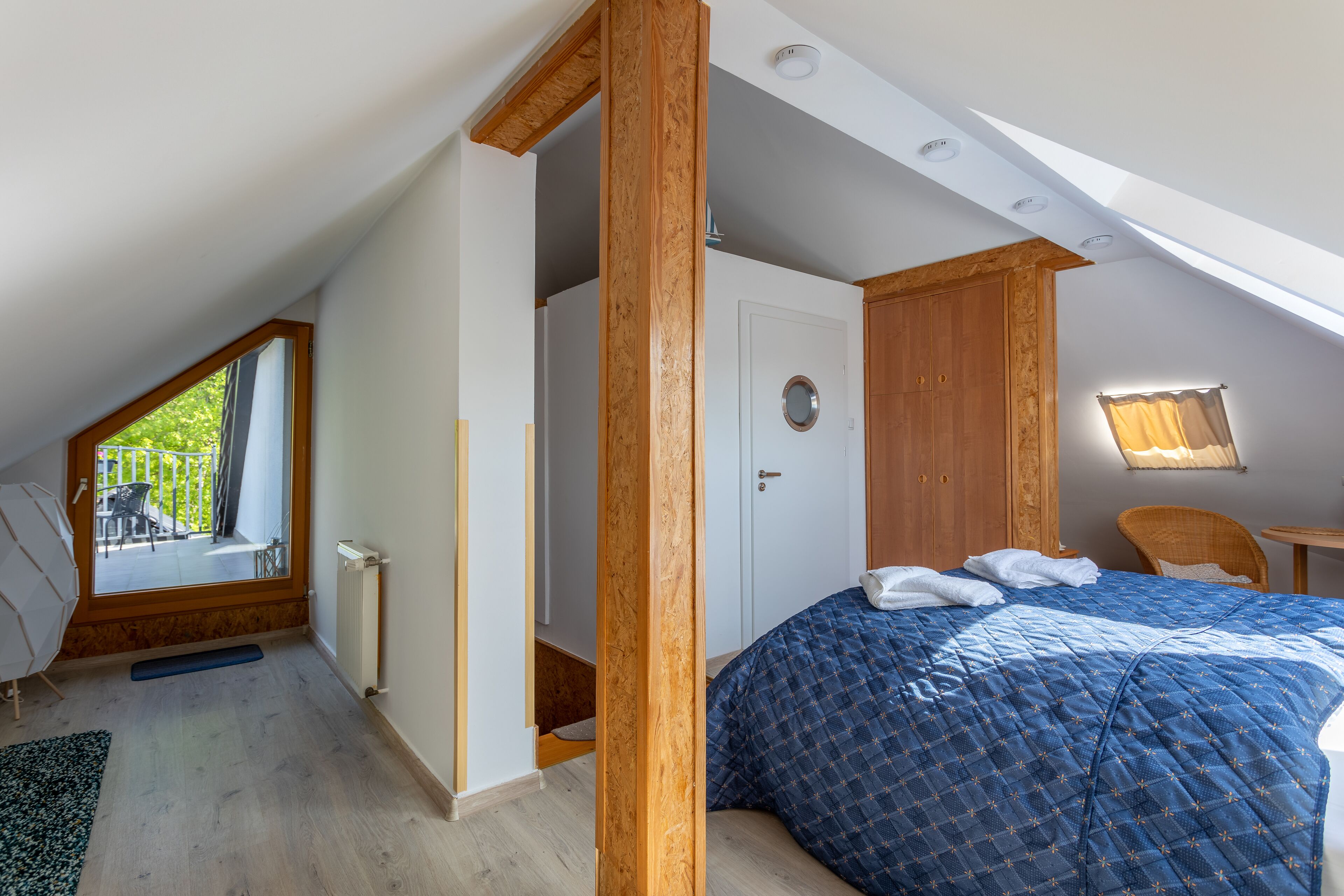 Ein Schlafzimmer mit schrägverlaufender Decke, einem Bett mit blauer Steudecke, einer Holztür und Blick auf einen Außenbalkon.